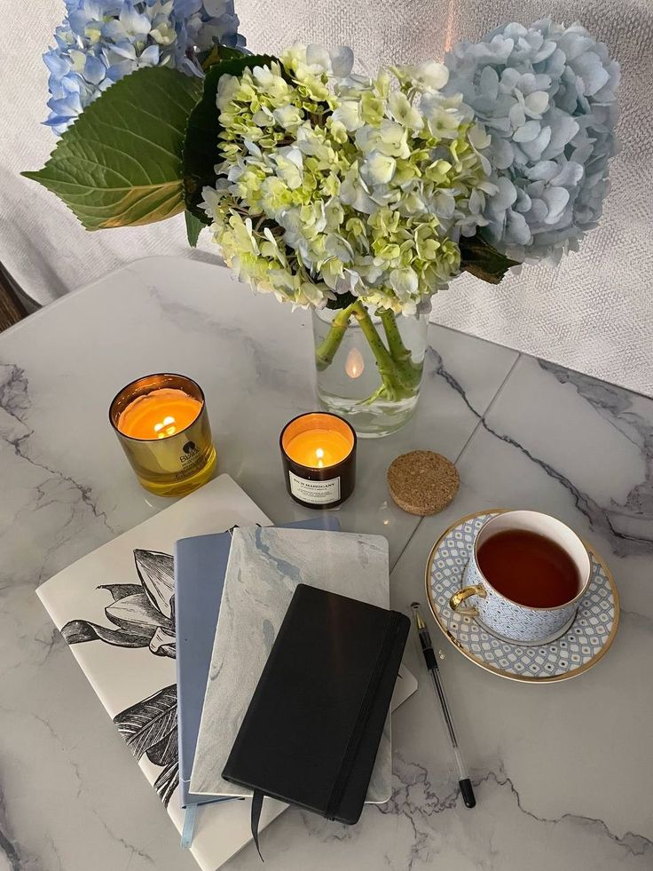 Cozy workspace with candles, notebooks, hydrangeas, and a cup of tea on a marble desk.