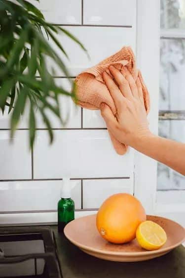Hand cleaning kitchen tiles with a cloth, beside a plate with citrus fruits.