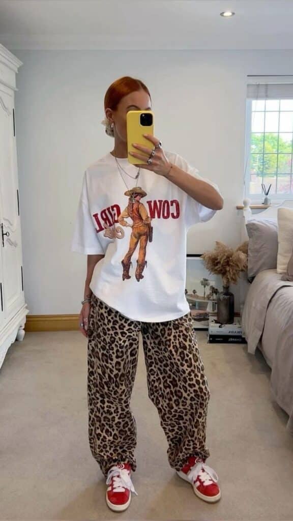 Person posing in mirror wearing cowboy-themed shirt, leopard print pants, and red sneakers.