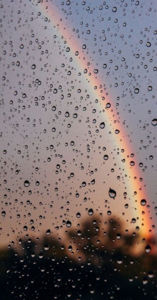 Raindrops on window with vibrant rainbow in the background at sunset.