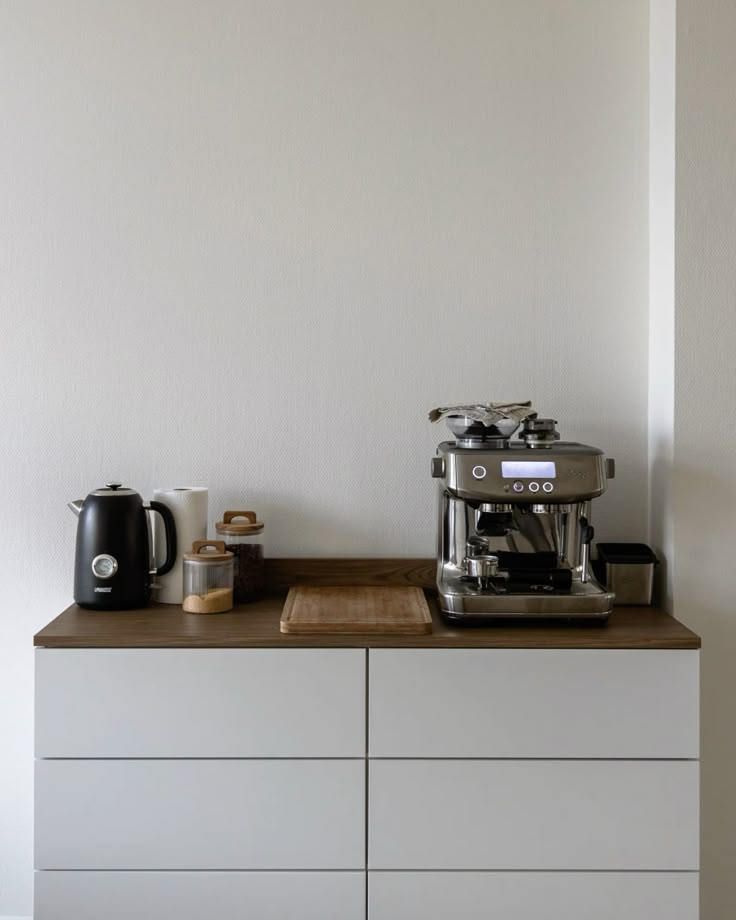 Modern kitchen counter with espresso machine, kettle, and wooden cutting board on a sleek cabinet setup.