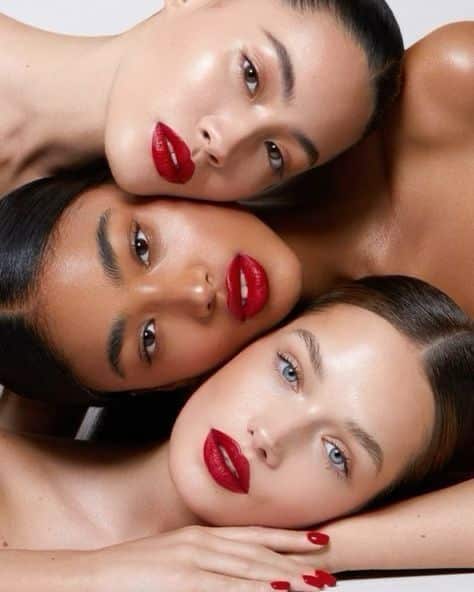 Three women with bold red lips and sleek hair posing closely in a beauty photoshoot.