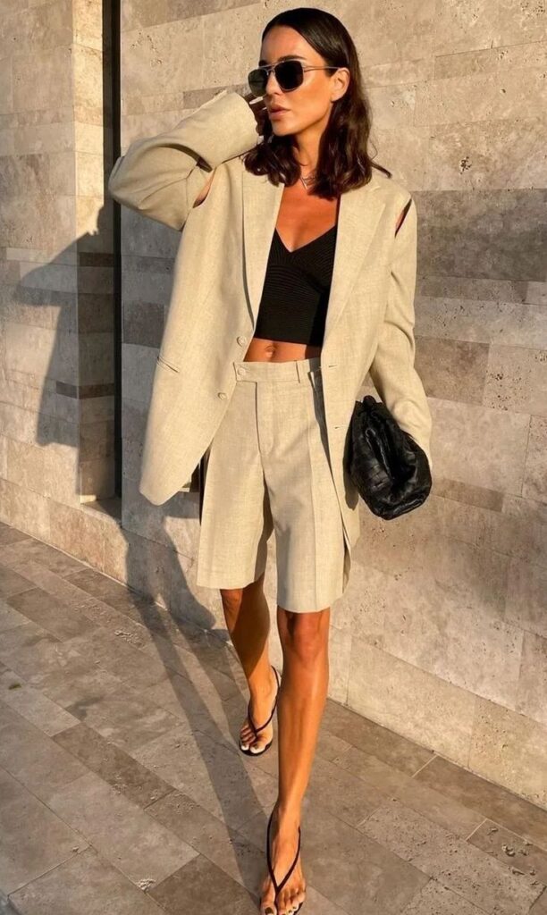 Stylish woman in beige blazer and shorts, black top, sunglasses, and sandals, walking confidently with a black clutch.