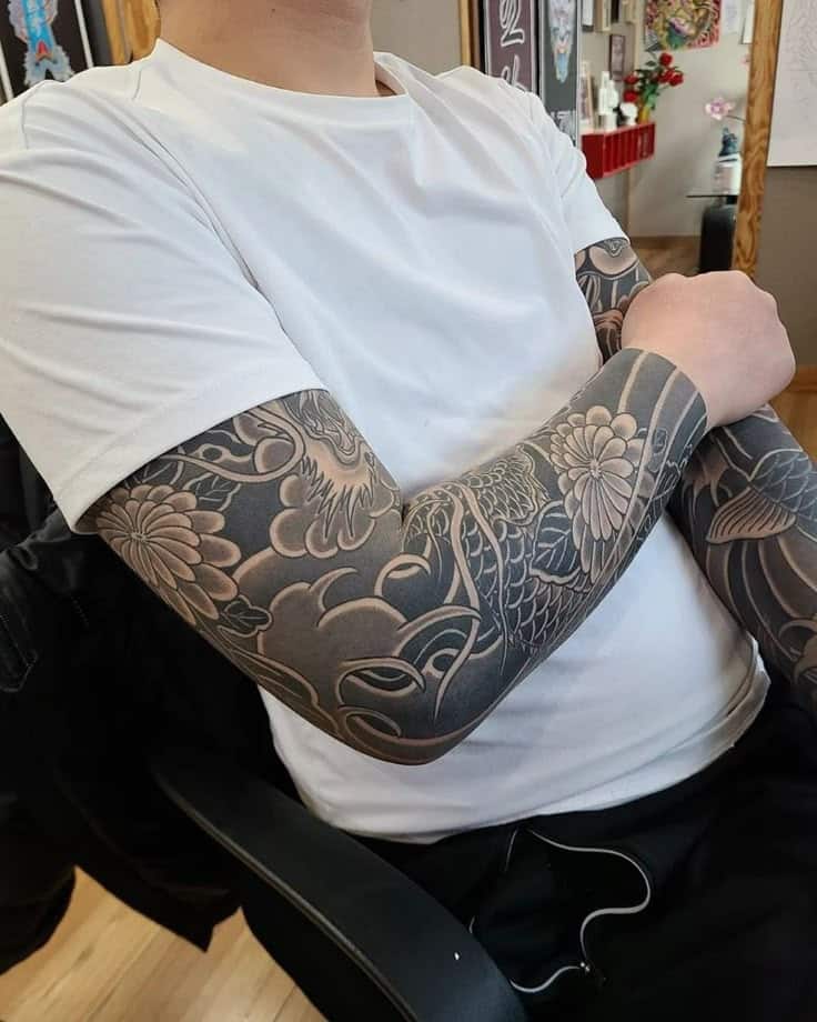 Person in white shirt with detailed black and gray sleeve tattoos sitting in a chair.