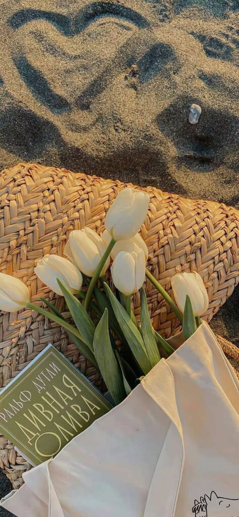 Beach scene with tulips in a woven bag, book, and heart drawn in sand. Cozy summer vibe.