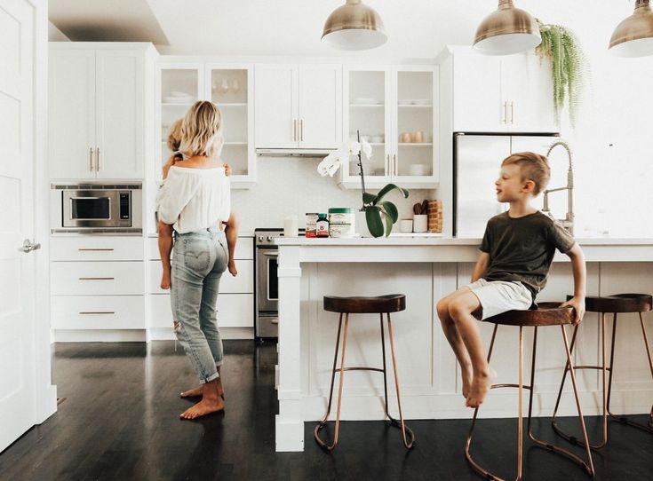 Modern kitchen with a woman holding a child, boy sitting on counter stool, white cabinets, and pendant lighting.
