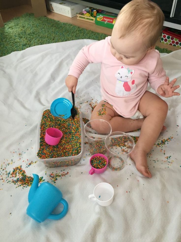 Baby playing with colorful sensory rice and cups on blanket.