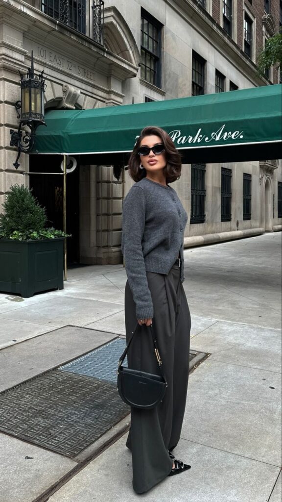 Fashionable woman in gray sweater and sunglasses, holding a black bag on a city street.
