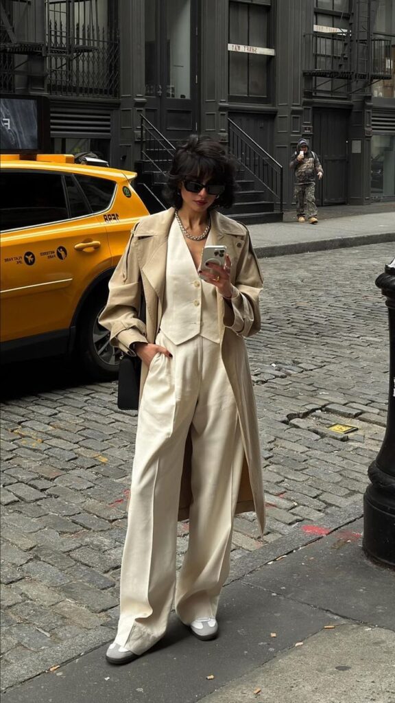 Stylish woman in chic beige outfit and trench coat checks phone on a cobblestone street near a New York taxi.