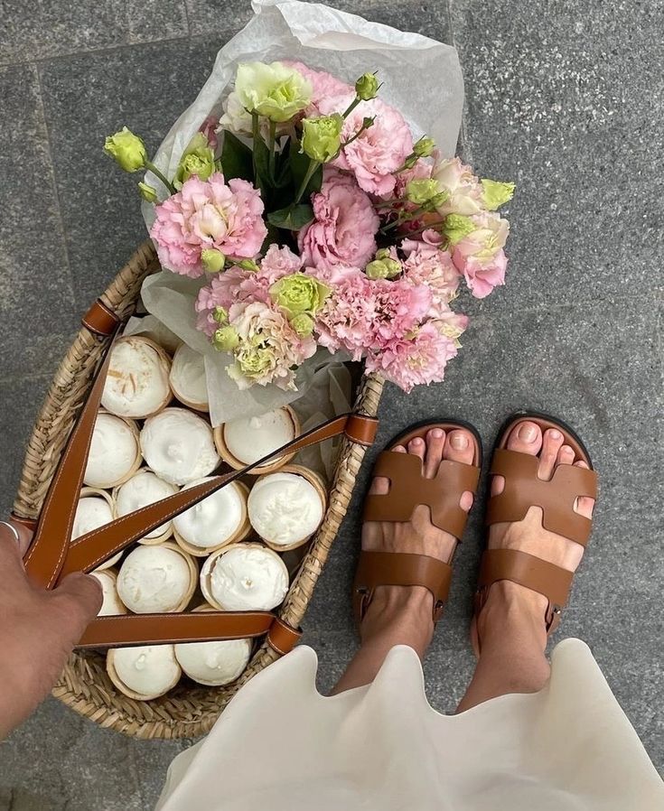 Wicker basket with pink flowers and cupcakes, person in brown sandals standing on a gray floor.
