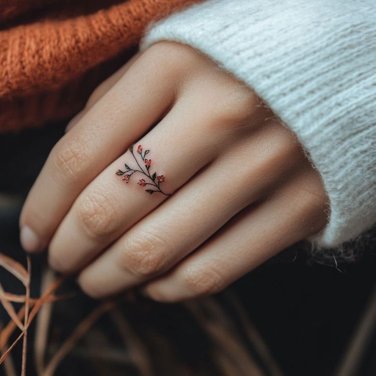 Delicate floral branch tattoo on finger, wearing cozy sweater. Minimalist hand ink design for artistic expression.