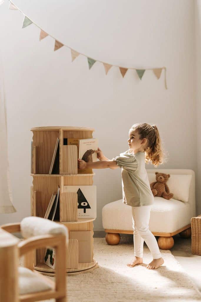 Young child reaches for book on wooden shelf in cozy, sunlit room with toy bear and bunting.