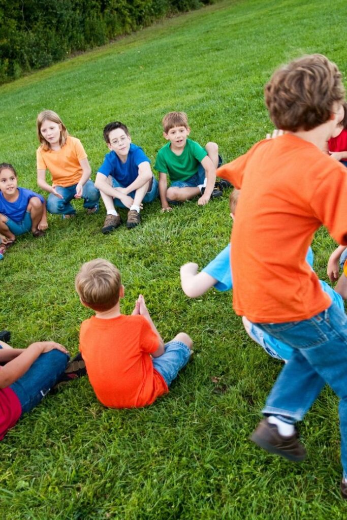 Children playing a game in a circle on green grass, wearing colorful t-shirts, enjoying outdoor fun.