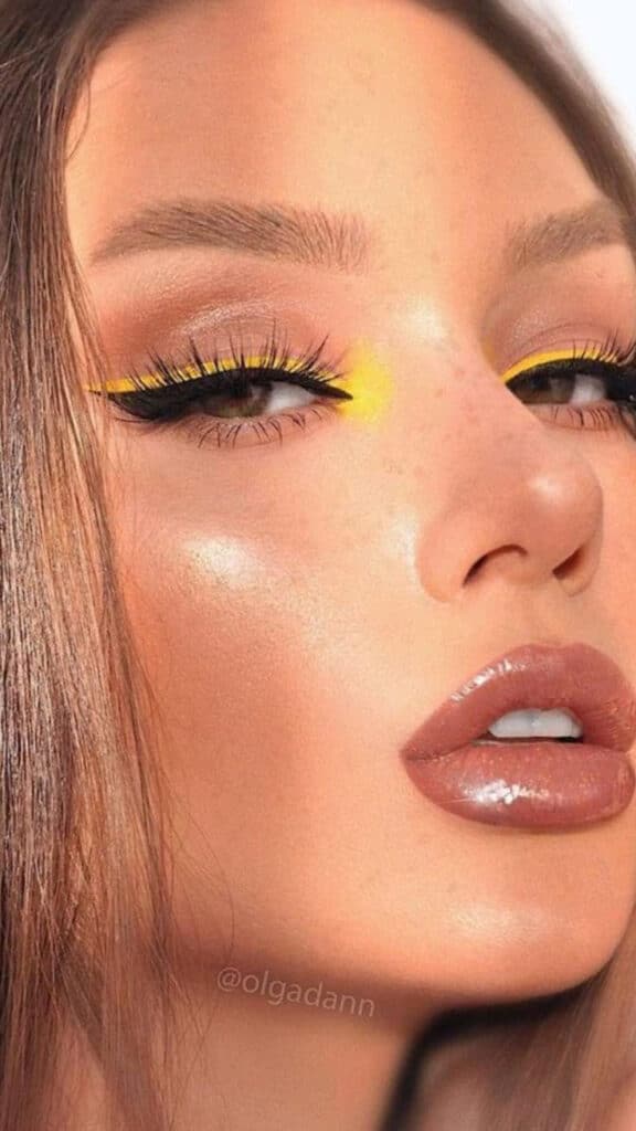 Close-up of a woman's face with bold yellow eye makeup and glossy lips, highlighting beauty trends.
