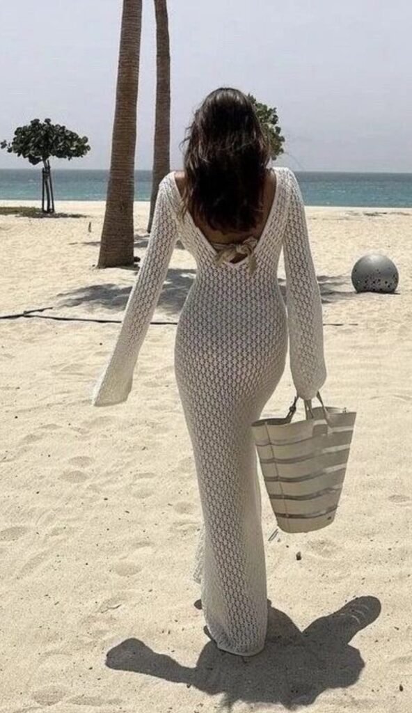 Woman in white crochet dress walking on sandy beach carrying a striped bag, with palm trees and ocean in background.