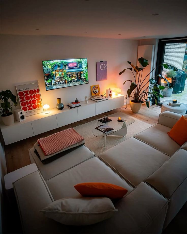 Modern living room with beige sectional sofa, wall-mounted TV, plants, and stylish decor. Cozy ambiance for relaxation.