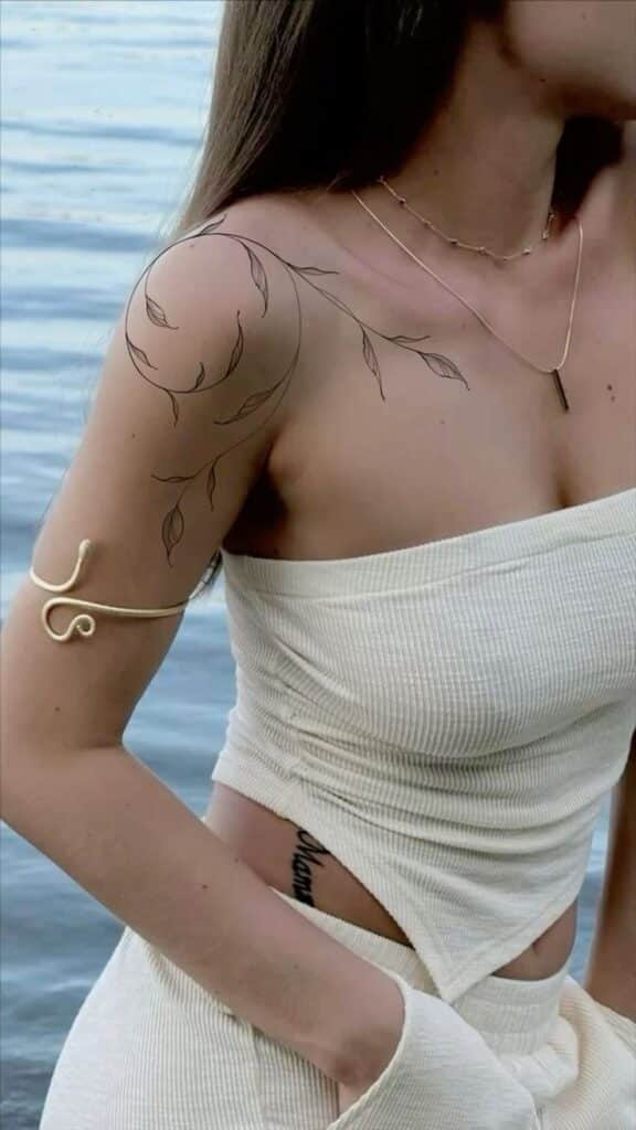 Woman with leaf tattoo and arm cuff in white outfit by water.