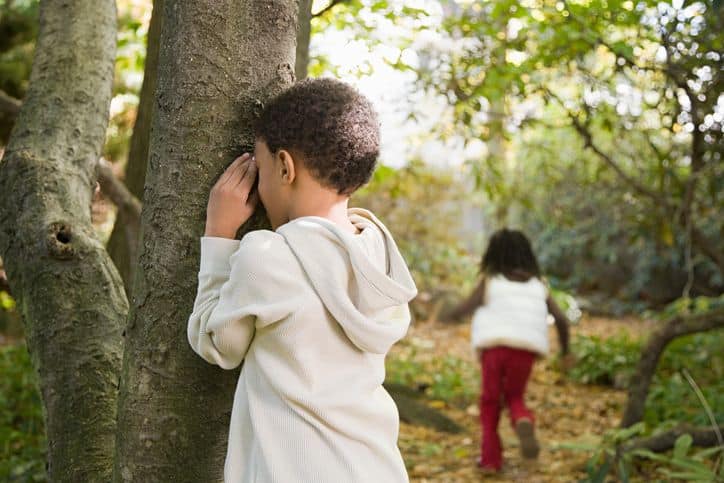 Children playing hide and seek in a sunlit forest, one counting by a tree while the other runs away.