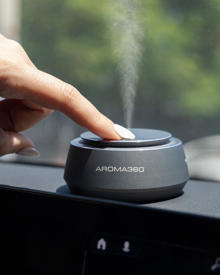 Finger pressing Aroma360 diffuser, releasing vapor in a car, enhancing interior with aromatic scent therapy.