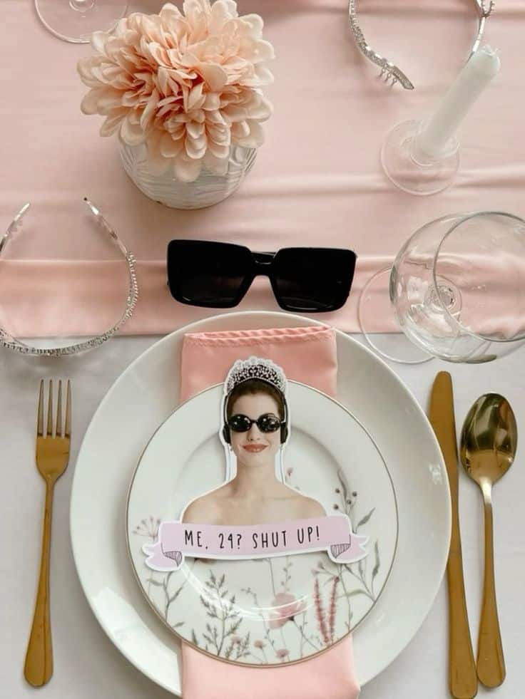 Elegant table setting with floral plate, humorous cutout, pink decor, gold cutlery, and stylish sunglasses.