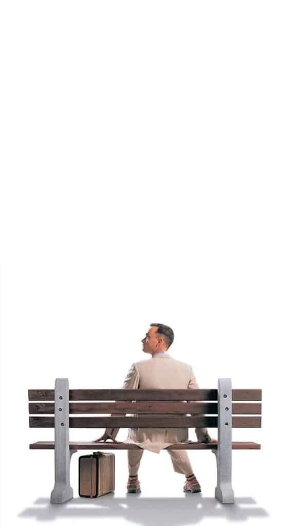 Man in a suit sitting on a bench with a suitcase, looking thoughtfully upward against a white background.