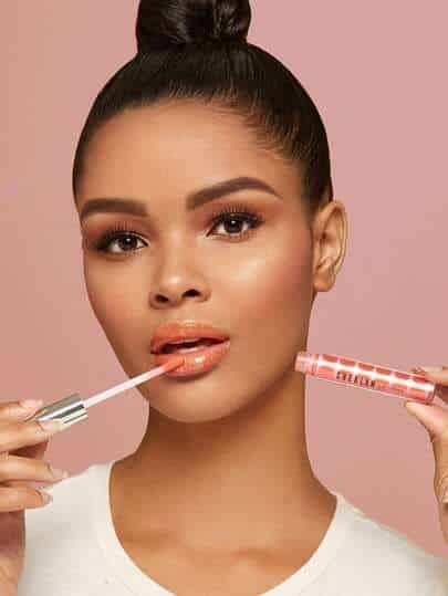 Woman applying glossy peach lip gloss on pink background, showcasing vibrant makeup look.