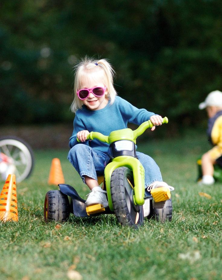 Toddler in pink sunglasses rides a toy green bike on grass, navigating orange cones with joy.
