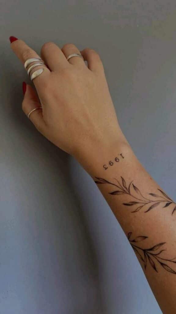 Arm with 1993 tattoo and leaf design, hand adorned with rings, showcasing minimalistic style against a gray background.