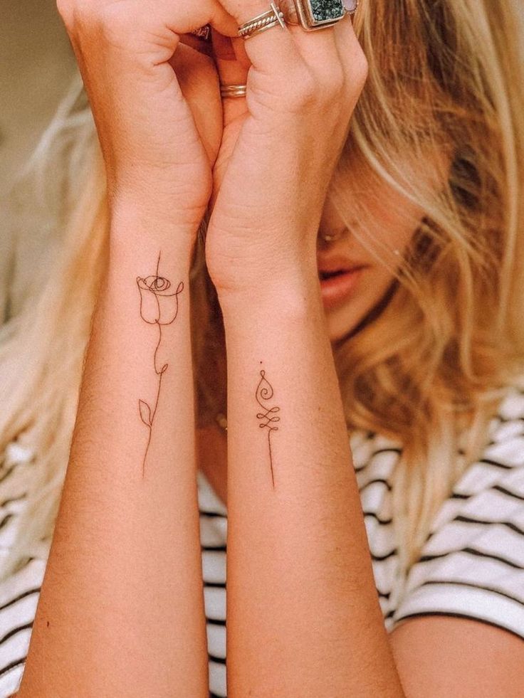 Minimalist line art tattoos of a flower and abstract design on forearms, woman wearing striped shirt.