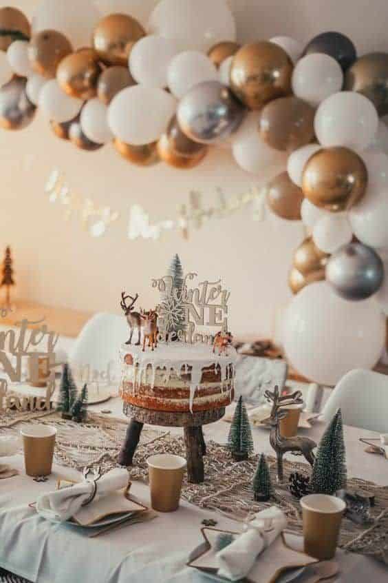Festive winter-themed birthday party table with cake, balloons, and decorations in gold, silver, and white.