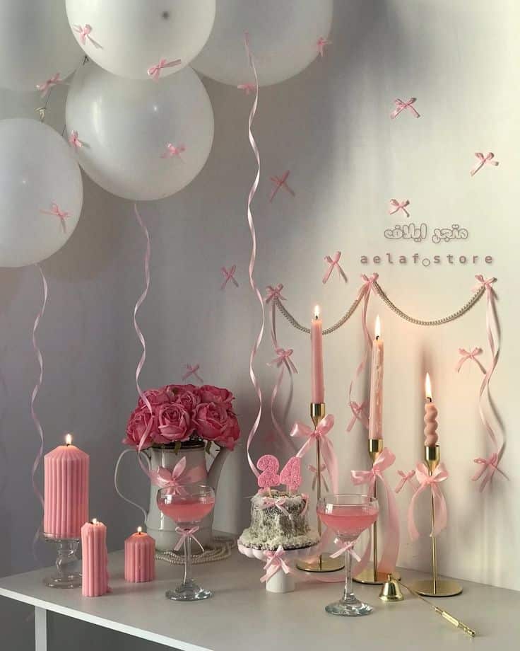 Elegant pink party setup with balloons, ribbons, flowers, candles, and drinks on a white table for a festive celebration.