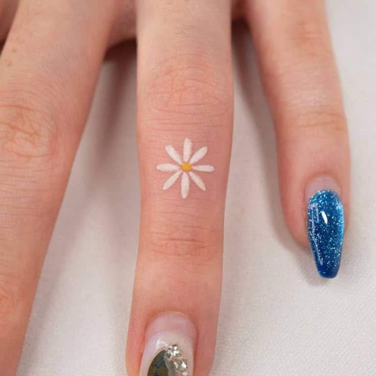 Minimalist daisy tattoo on finger with blue glitter nail design.