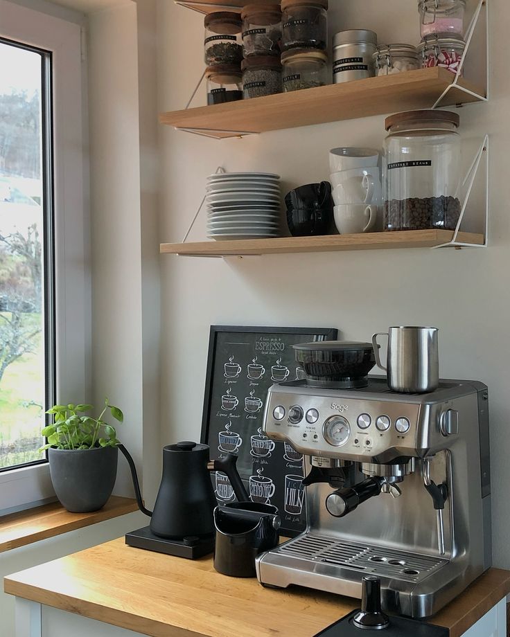 Modern kitchen coffee station with espresso machine, mugs, and shelves of coffee jars by a window.
