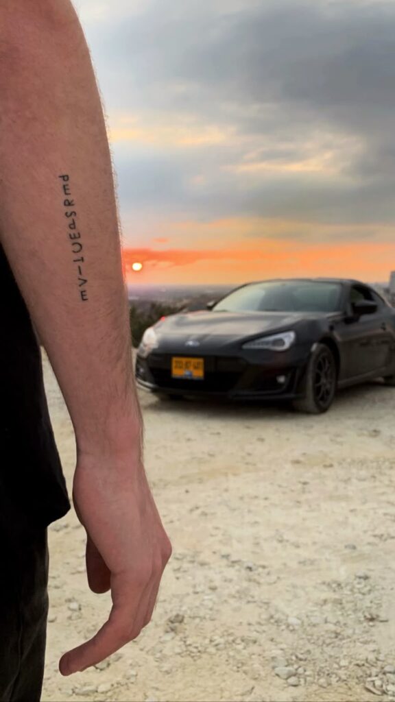 Arm with tattoo in foreground, black sports car and sunset in the background on a gravel road.