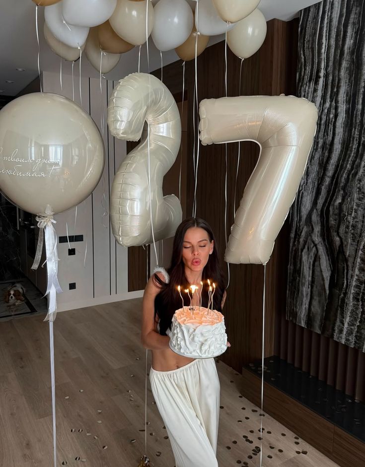 Woman celebrating 27th birthday with cake and balloons indoors.