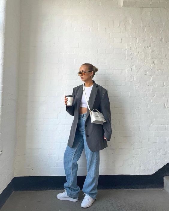 Fashionable woman in oversized blazer, jeans, and sunglasses holding coffee. Casual chic street style with a white crossbody bag.