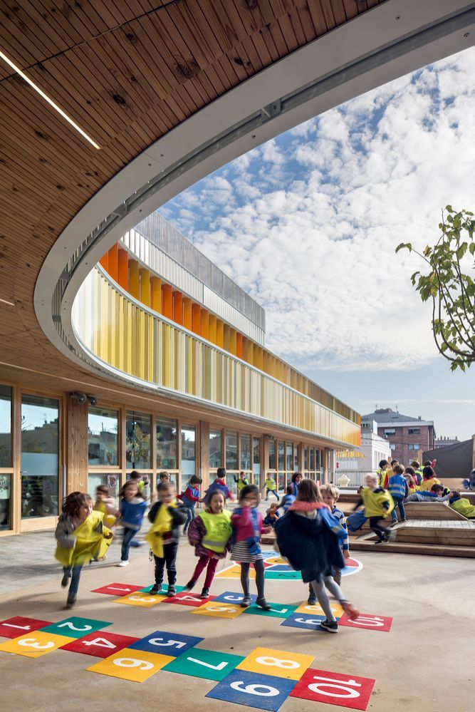 Children playing hopscotch on a colorful playground at a modern, curved architectural school building under a bright sky.