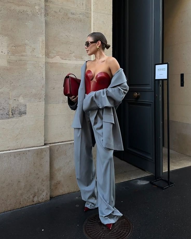 Fashionable woman wearing gray suit, red corset, and sunglasses, holding a bag outdoors against a building background.