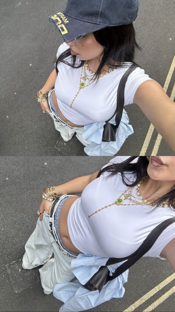 Fashionable woman in casual outfit with white crop top, blue cap, layered jewelry, and baggy pants on urban street.