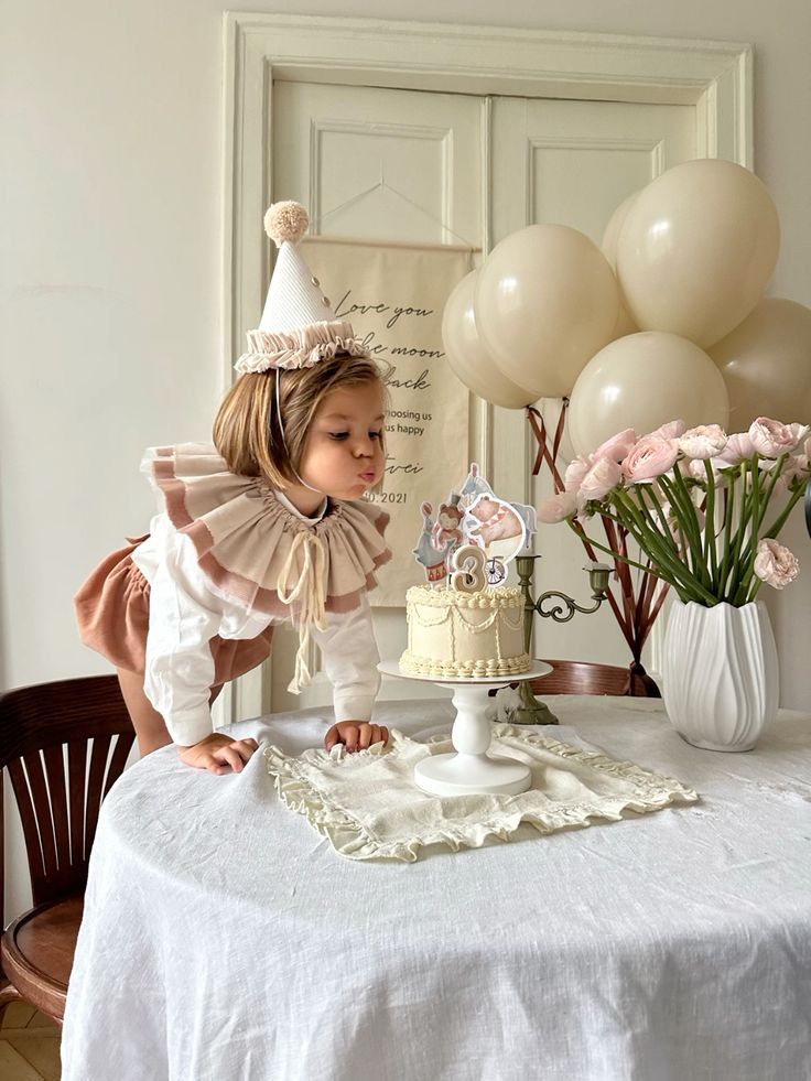 Child in a party hat blows out candles on a cake at a birthday celebration surrounded by balloons and flowers.