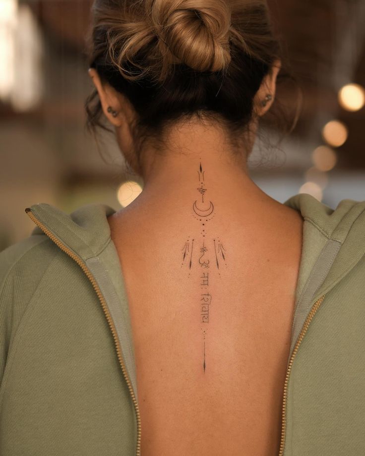 Back tattoo design with crescent moon and symbols, woman with hair bun, wearing green hoodie, creative body art.