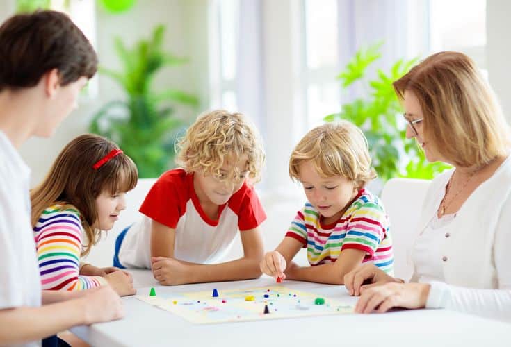 Children and adult playing a board game together, sitting around a table in a bright room with green plants.