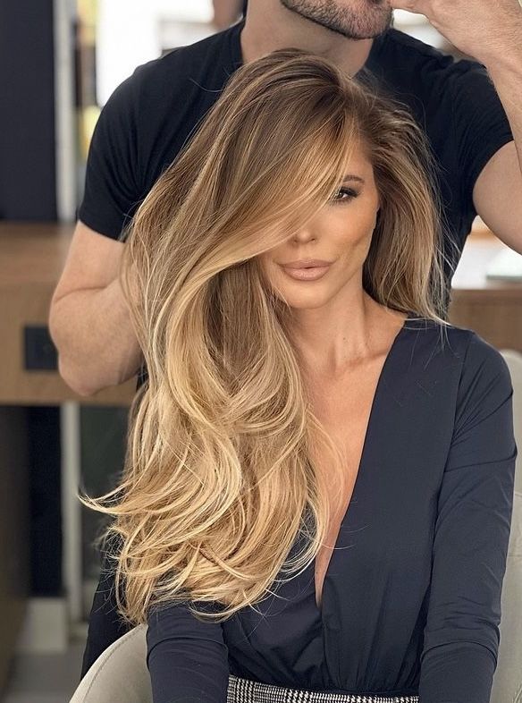Woman getting styled with long, voluminous blonde hair in a salon, wearing a dark top.