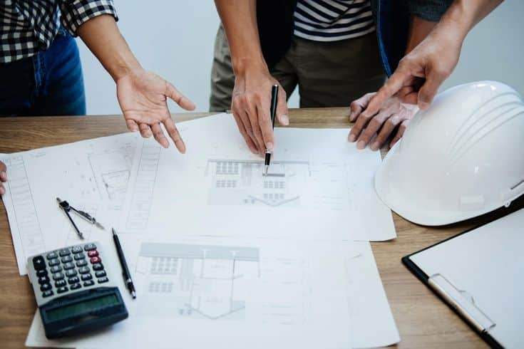 5 Questions To Ask Before Hiring A Remodeling Contractor People discussing architectural plans on a table with a hard hat, calculator, and clipboard. | Sky Rye Design People discussing architectural plans on a table with a hard hat, calculator, and clipboard.