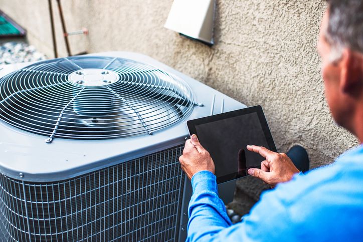 Technician using tablet to check outdoor AC unit functionality against a wall.