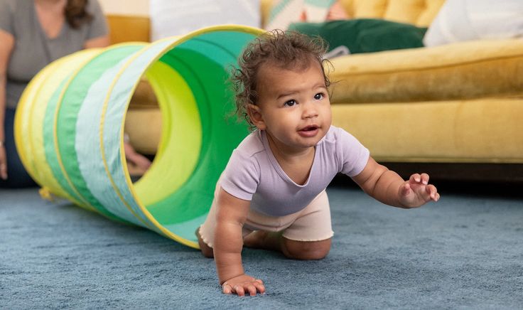 Baby crawling through play tunnel on blue carpet in living room with yellow couch.