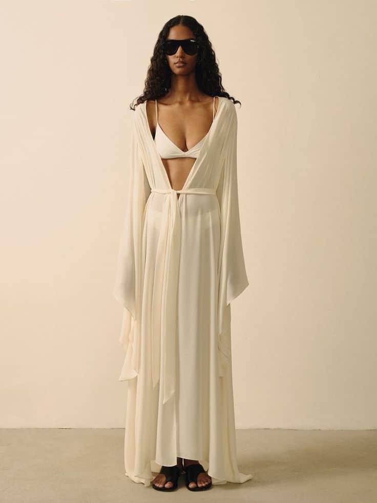 Woman in elegant white dress and sunglasses, standing against neutral background, showcasing fashion style.
