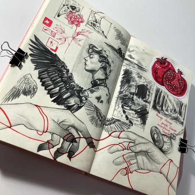 Sketchbook art with graphite sketches of wings, a figure, and hands, detailed with red highlights and pomegranate sticker.