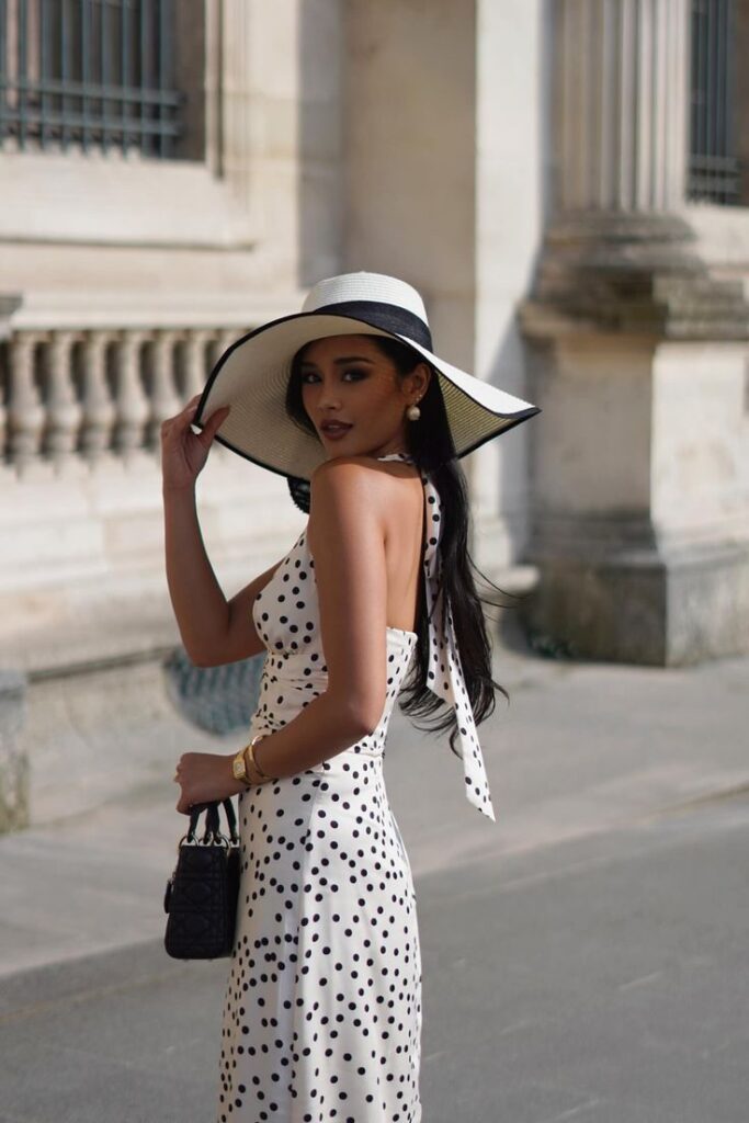 Woman in polka dot dress and wide-brimmed hat walking outside a historic building.