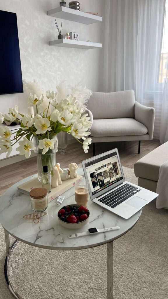 Cozy living room with laptop, flowers, and fruit bowl on marble table. Modern home office setup with natural light.